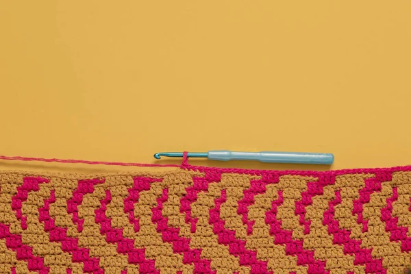 Yellow pink crochet fabric with blue crochet hook on a yellow background. Top view. Copy space.