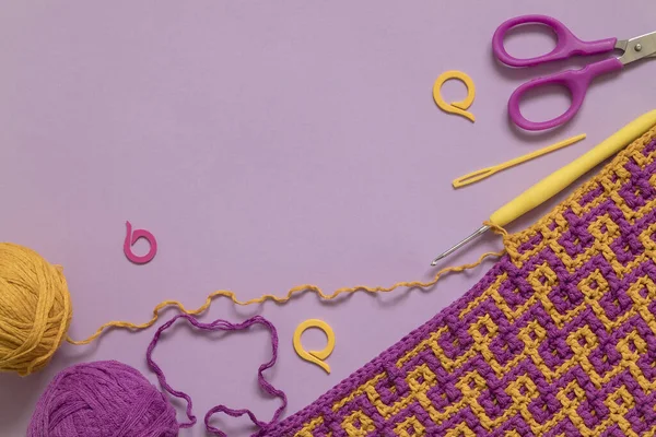 Crochet fabric with abstract pattern, crochet hook, scissors, knitting markers and yarns on a purple background. Copy space.