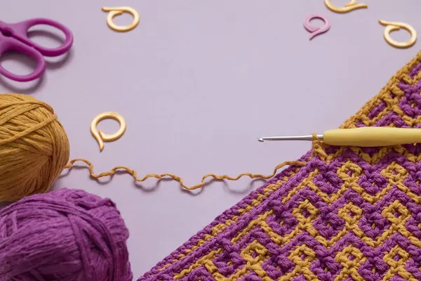 Workspace with crochet fabric and crochet tools on a purple background. Copy space.