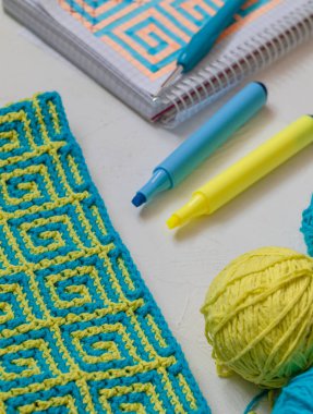 Mozaik desenli sarı mavi örgü kumaş. Tığ işi kumaş, iplik topları, tığ işi kanca, defter ve kalemlerle dolu bir yer. Ev kavramında örgü öreriz..