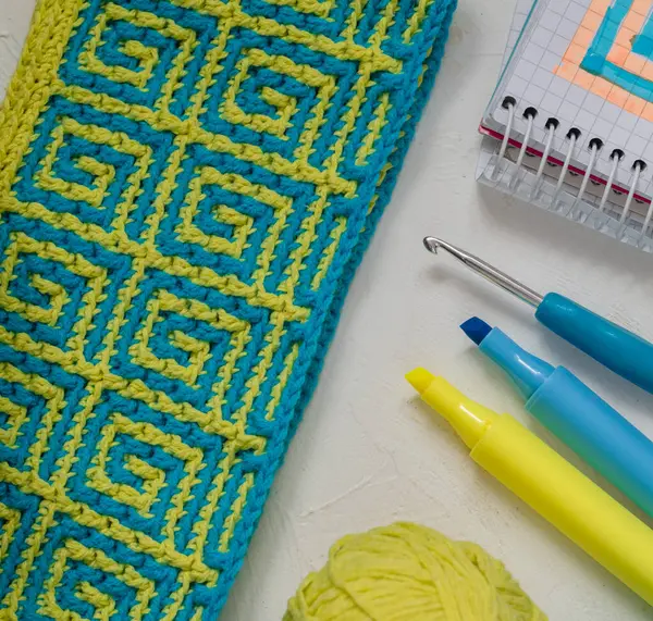 Mozaik desenli sarı mavi örgü kumaş. Yaklaş. Tığ işi kumaş, iplik topları, tığ işi kanca, defter ve kalemlerle dolu bir yer..