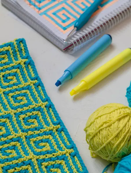 Mozaik desenli sarı mavi örgü kumaş. Tığ işi kumaş, iplik topları, tığ işi kanca, defter ve kalemlerle dolu bir yer. Ev kavramında örgü öreriz..