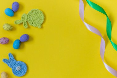 Easter composition with crochet bunnies, colorful eggs and satin ribbon on a yellow background. Top view. Copy space.