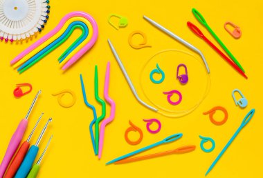 Sarı arka planda örgü aletleri. Tığ işi kancalar, plastik örgü iğneleri, işaretler, iğneler, eğri iğneler. Düz yatıyordu. Üst görünüm.