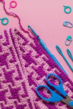 Pembe örgü örme mozaik kumaş, mavi örgü kancası makası. Pembe arka planda iğneler ve işaretler var. Boşluğu kopyala.