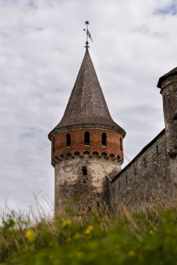 Ortaçağ kalesinin kulesinde konik bir çatı ve taş duvarlar, yeşil ve bulutlardan oluşan bir zemin oluşturarak mimari miras ve doğayı ön plana çıkarıyor.