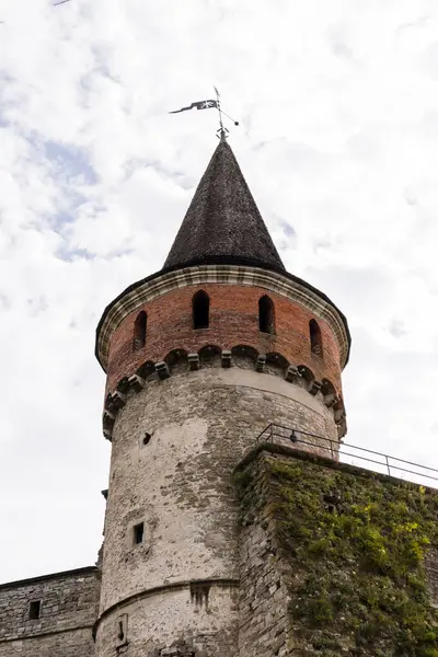 Bir ortaçağ kalesinin kulesinde taş duvarlar ve yeşil ile çevrili konik bir çatı bulunur ve dramatik bir gökyüzü altında mimari güzellik ve tarihi ön plana çıkarır.
