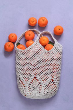 Mor arka planda taze organik mandalinalarla dolu beyaz pamuklu ağ torbası. Ananas desenli tığ işi çanta. Çevre dostu alışveriş. Boşluğu kopyala.