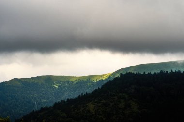 Yeşil bir dağ sırtı üzerinde kara, uğursuz bir fırtına bulutu asılı dramatik bir manzara.
