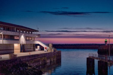 Solunda bir bina olan bir limanın alacakaranlık manzarası ve suya uzanan bir iskele. Gökyüzü maviden turuncuya dönüşür ve uzaklıktan yeşil ışık görünür..