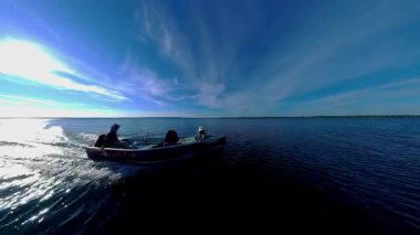 Kara siluetiyle altın saat batımında süzülen balıkçı tekneleriyle turistler. Balık tutmak için en iyi zaman. Tekneden güneşin battığı göl manzarası. Akşam eğlencesinin sinematik görüntüsü.