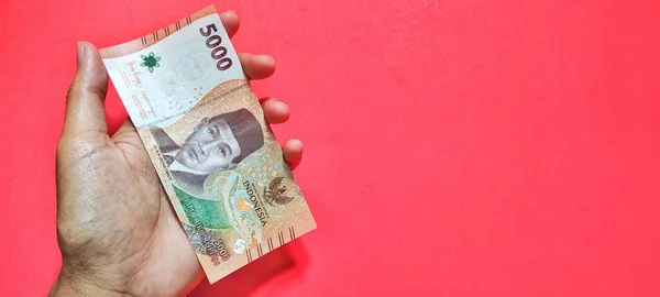 A man holding a new Indonesian 5,000 rupiah banknote issued in 2022. Rupiah currency concept isolated on a red background