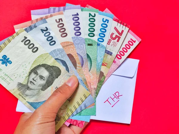 2022 'de 1000 Rupi' den 100.000 Rupi 'ye kadar yeni banknotlar tutan adam. Üzerinde THR yazan beyaz bir zarf olan Endonezya rupia parası. Tunjangan Hari Raya kavramı kırmızı arkaplanda izole edildi