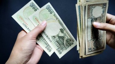 Rp10 'dan Rp500' e kadar olan eski rupiah banknotlarını sayarken bir kişinin eli görülüyor, siyah bir arka planda izole edilmiş. Yakın çekim görünümü