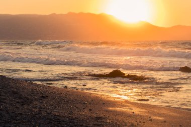 Dalgalar Yunanistan 'da cennet gibi bir sahile vuruyor. Denizin dalgalı olduğu bir yaz günü. Arka planda güneş dağların ardında batar.