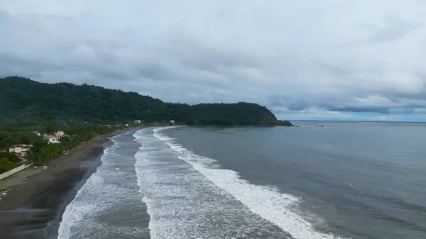 Geniş bir hava manzarası oldukça bulutlu bir günde güzel bir plajı gösteriyor, dalgalar yavaşça kıyıya vuruyor.