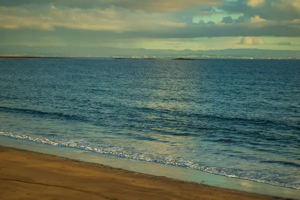 Bulutlu gökyüzünün altında sakin deniz dalgalarıyla huzurlu kumlu sahil