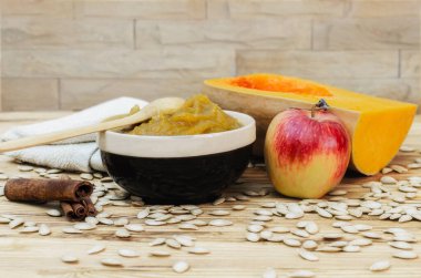 Ahşap bir masada balkabağı lapası ve dekoratif bir duvarın arka planında dağınık elma ve tarçın tohumları..