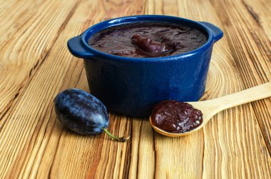 Ahşap bir arka planda, mavi bir seramik kapta erik reçeli, yanında da ahşap bir kaşık, taze erikler.