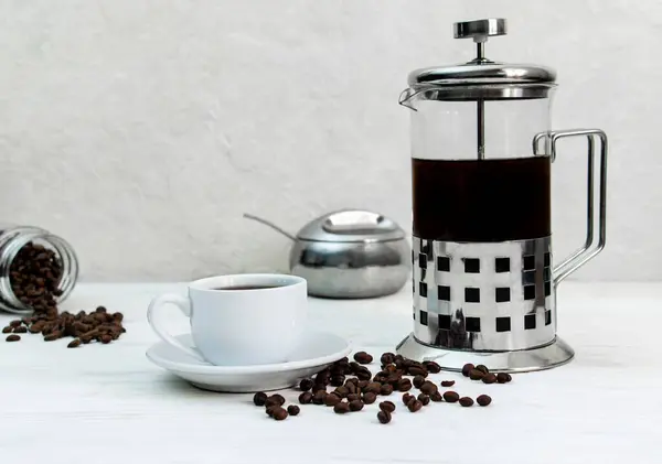 White coffee mug on a white plate with a french press.Scattered coffee beans on a white wooden surface