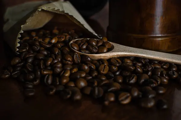 stock image Freshly roasted coffee beans spilling from a partially opened brown paper bag onto a wooden surface. A wooden spoon lies among the grains