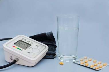 Kan basıncı monitörüne yakın çekim, bir bardak su ve hipertansiyon tedavisini sembolize eden su toplayan ilaçlar..