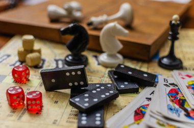 Çeşitli klasik masa oyunlarının yakın plan natürmortu. Sahnede domino taşları, kırmızı zarlar, iskambil kağıtları ve satranç taşları da dahil olmak üzere boş zaman ve hobi eşyaları koleksiyonu yer alıyor..