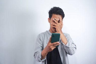 Beyaz arka planda izole edilmiş cep telefonuna bakarken bir gözünü kapatan korkmuş Asyalı adamın yakın plan portresi.