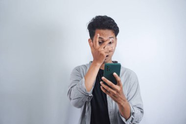 Beyaz arka planda izole edilmiş cep telefonuna bakarken bir gözünü kapatan korkmuş Asyalı adamın yakın plan portresi.