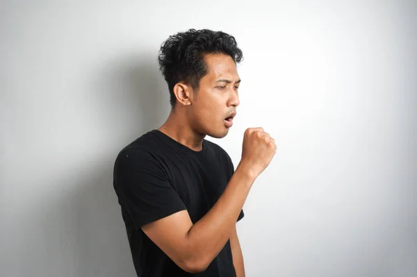 young man feeling ill with a sore throat and flu symptoms, coughing with mouth covered against white wall