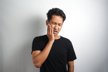 Portre genç adam yanağa dokunur. Güçlü bir diş ağrısı ve gri arka planda günlük beyaz tişört giydiği için acı çekiyor. Diş ağrısını hisset