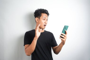 Asyalı adamın yüzü, gri arka planda akıllı telefonda gördüklerini şok etti. Endonezyalı adam siyah gömlek giyer. Gri arka plan..