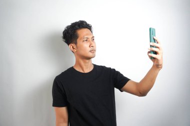 Siyah tişört giyen ve beyaz arka planda yalnız poz veren mutlu Asyalı genç bir adam resmi. Telefonla selfie çekin..