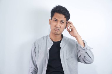 Tişörtlü genç adamın şüpheleri var ve kafası karman çorman, beyaz arka planda kaşınırken yüz ifadesi karışık.