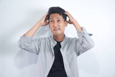 Tişörtlü genç adamın şüpheleri var ve kafası karman çorman, beyaz arka planda kaşınırken yüz ifadesi karışık.