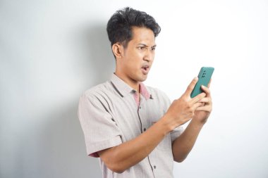 Amazed shocked asian guy holding smartphone in his hand, looking at the phone in surprise, stunned facial expression, stands on isolated white background