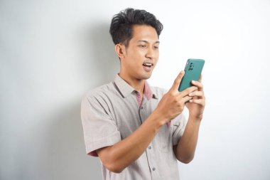 Amazed shocked asian guy holding smartphone in his hand, looking at the phone in surprise, stunned facial expression, stands on isolated white background