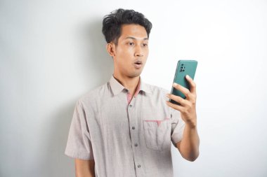 Amazed shocked asian guy holding smartphone in his hand, looking at the phone in surprise, stunned facial expression, stands on isolated white background