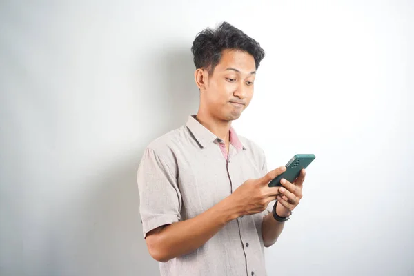 Thoughtful handsome asian man standing on white background with smartphone in hand, looking and thinking wearing shirt. Isolated. Copy space