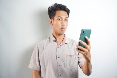 Amazed shocked asian guy holding smartphone in his hand, looking at the phone in surprise, stunned facial expression, stands on isolated white background