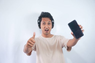 Half length shot of optimistic man holds smartphone with mock up screen, listens favourite soundtrack in headphones, wears casual cream t shirt, stands indoor. People and technology concept.