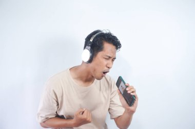 Energized man sings to music, moves actively, wears headphones and casual t shirt, poses against white background, keeps mouth widely opened, enjoys life, isolated on white studio wall