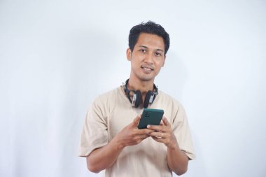 Handsome man student with headphones, holding Cellphone, reading message on mobile phone, standing against light white background