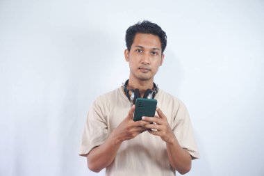 Handsome man student with headphones, holding Cellphone, reading message on mobile phone, standing against light white background
