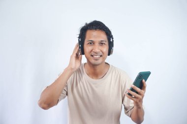 Young smiling fun cool man of Asian ethnicity 20s wearing cream shirt headphones listen to music hold use mobile cell phone isolated on plain white background studio portrait