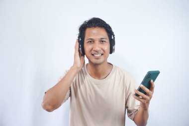 Young smiling fun cool man of Asian ethnicity 20s wearing cream shirt headphones listen to music hold use mobile cell phone isolated on plain white background studio portrait