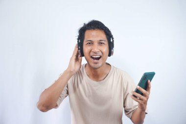 Young smiling fun cool man of Asian ethnicity 20s wearing cream shirt headphones listen to music hold use mobile cell phone isolated on plain white background studio portrait