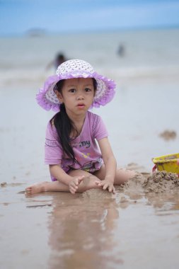 Balikpapan, 1 Mart 2023. Mor plaj şapkası ve tişörtü olan şirin küçük bir kız kumsalda kumsalda oynuyor. Tatil ve rahatlama. Yaz tatilinde sahilde aktif çocuk ve çocuk gelişimi..