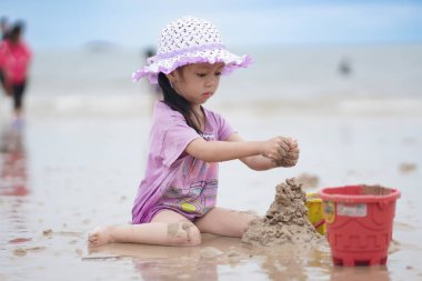 Balikpapan, 1 Mart 2023. Mor plaj şapkası ve tişörtü olan şirin küçük bir kız kumsalda kumsalda oynuyor. Tatil ve rahatlama. Yaz tatilinde sahilde aktif çocuk ve çocuk gelişimi..