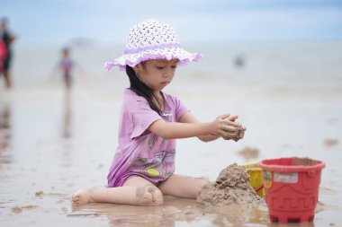 Balikpapan, 1 Mart 2023. Mor plaj şapkası ve tişörtü olan şirin küçük bir kız kumsalda kumsalda oynuyor. Tatil ve rahatlama. Yaz tatilinde sahilde aktif çocuk ve çocuk gelişimi..
